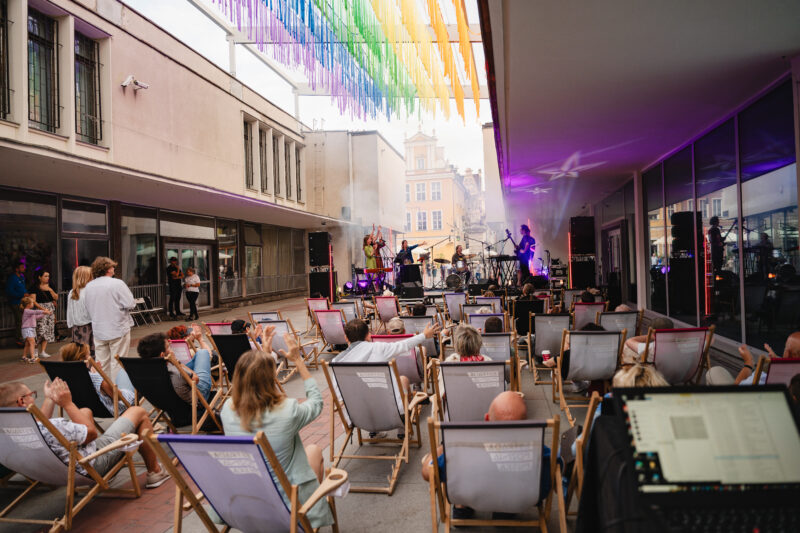 Koncert na starym runku w Poznaniu: Zespół na scenie, publiczność na leżakach, kolorowe dekoracje zawieszone między budynkami.