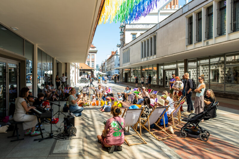 Koncert dla dzieci na Starym rynku: Muzycy oraz dzieci na leżakach. Kolorowe dekoracje między budynkami.