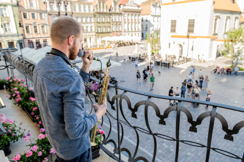 Koncert na świeżym powietrzu: Muzyk z mikrofonem oraz saksofonem na balkonie z widokiem na Stary Rynek w Poznaniu.