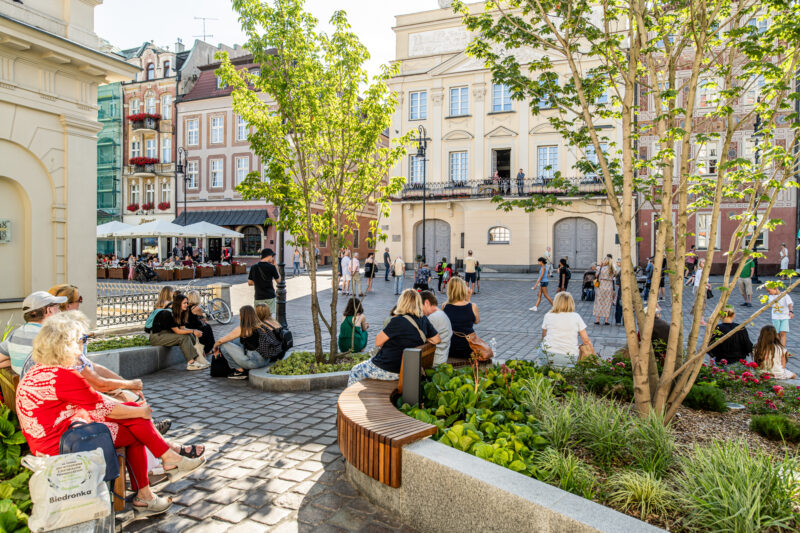 Publiczność koncertu na wolnym powietrzu, siedząca na ławkach otoczonych zielenią. W tle kolorowe kamienice.