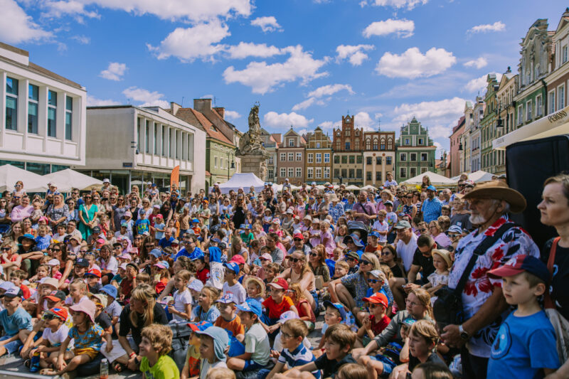 Tłum ludzi, głównie dzieci oświetlony słońcem i otoczony kolorowymi kamienicami na Starym Rynku w Poznaniu.