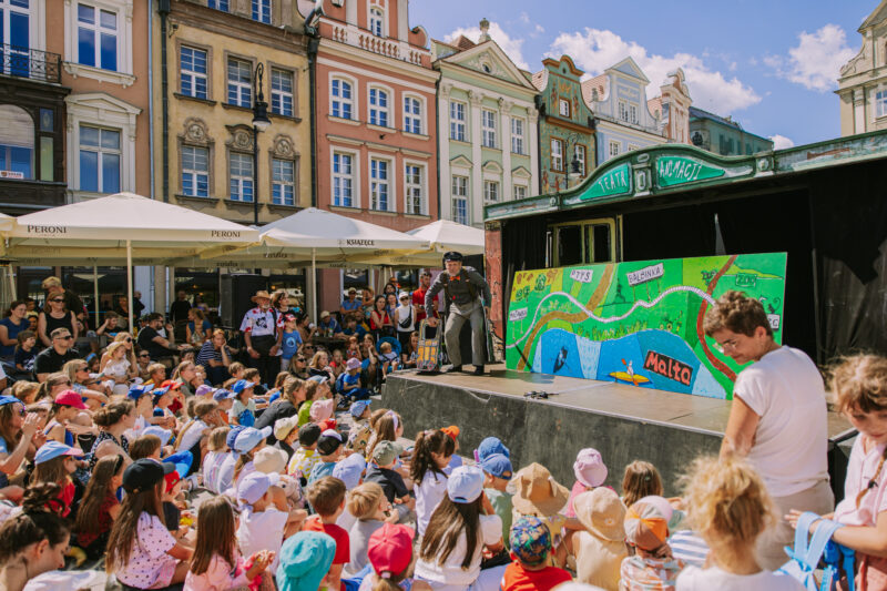 Spektakl dla dzieci na świeżym powietrzu, Stary rynek w Poznaniu. Na scenie starszy aktor oraz duża mapa. Wszystko na tle kolorowych kamienic.