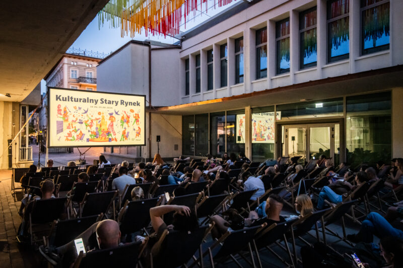 Kino plenerowe na Starym Rynku w Poznaniu: Publiczność na leżakach, na ekranie kolorowa identyfikacja wizualna Estrady Poznańskiej.